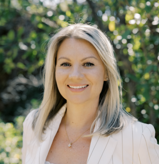 A woman with straight blonde hair, wearing a white blazer and a necklace, smiles outdoors in bright, natural light with green foliage in the background.
